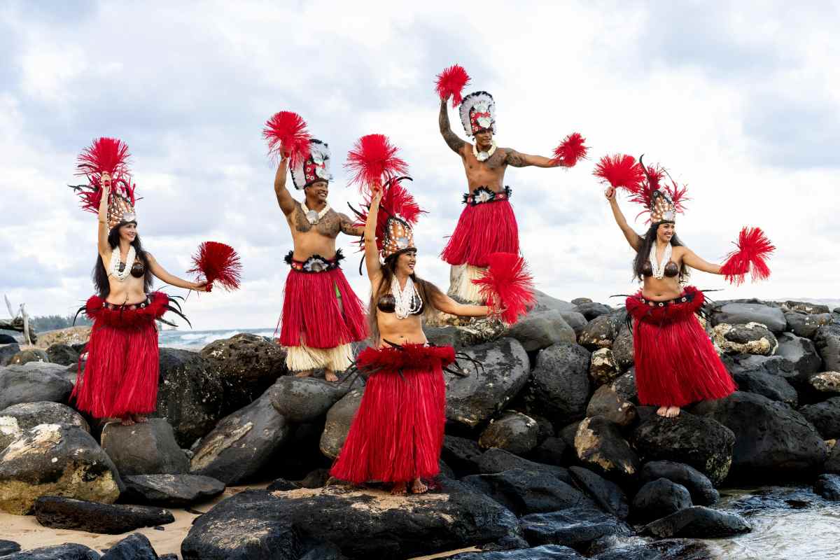 Five people in red skirts and feathered headpieces dance on rocks by the sea.